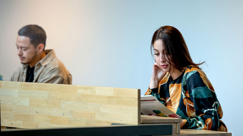 Female student reading at a high table, male student blurry in the background